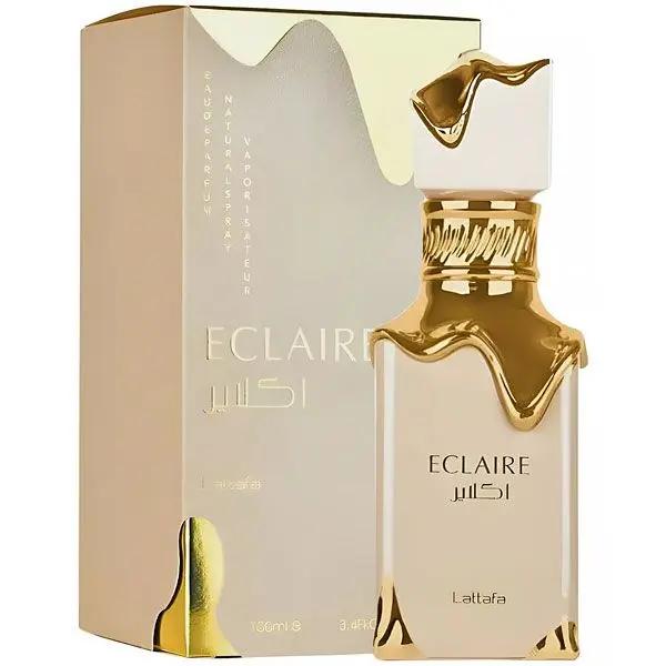 Un frasco de perfume Lattafa ECLAIRE dorado y crema de Royal Oud Perfumes, ideal como fragancia gourmand para mujer, presenta un tapón decorativo a juego y se exhibe delante de su elegante y brillante estuche de marca.